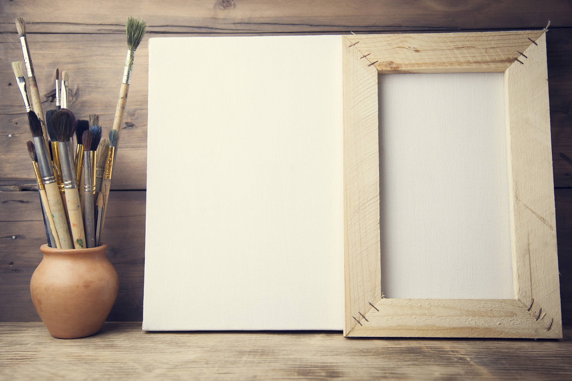 Art supplies include a pot of paintbrushes beside an empty canvas framed in wood, set against a rustic wooden background.