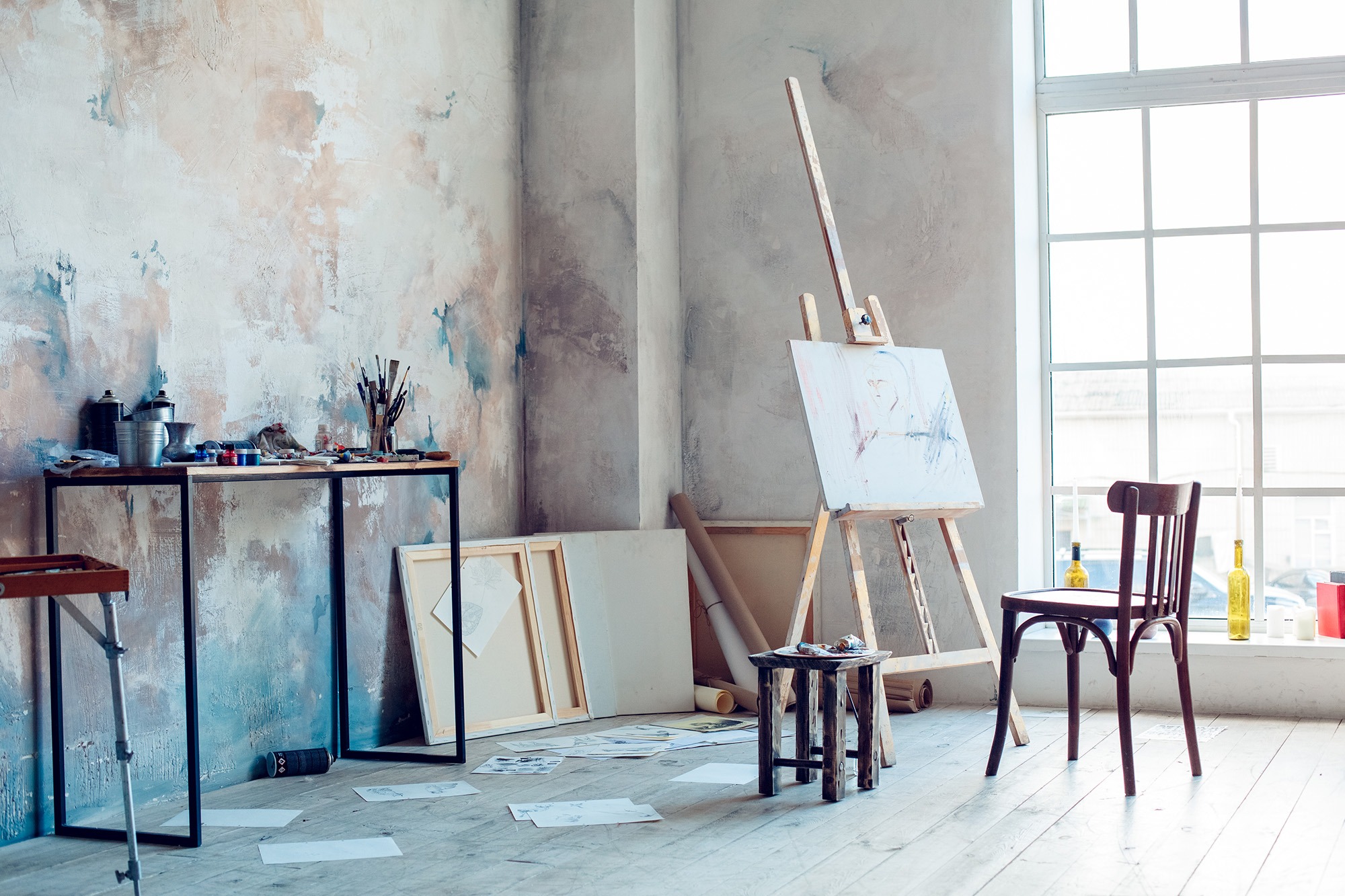 Sunlit art studio featuring a wooden easel with a canvas, a cluttered table with paint supplies, and sketches scattered on the floor.
